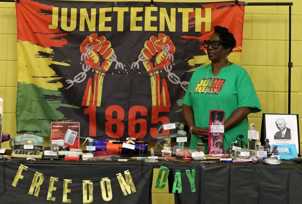 A woman in a green Juneteenth shirt stands in front of a Juneteenth banner with the number 1865, and a table with historical artifacts and images.