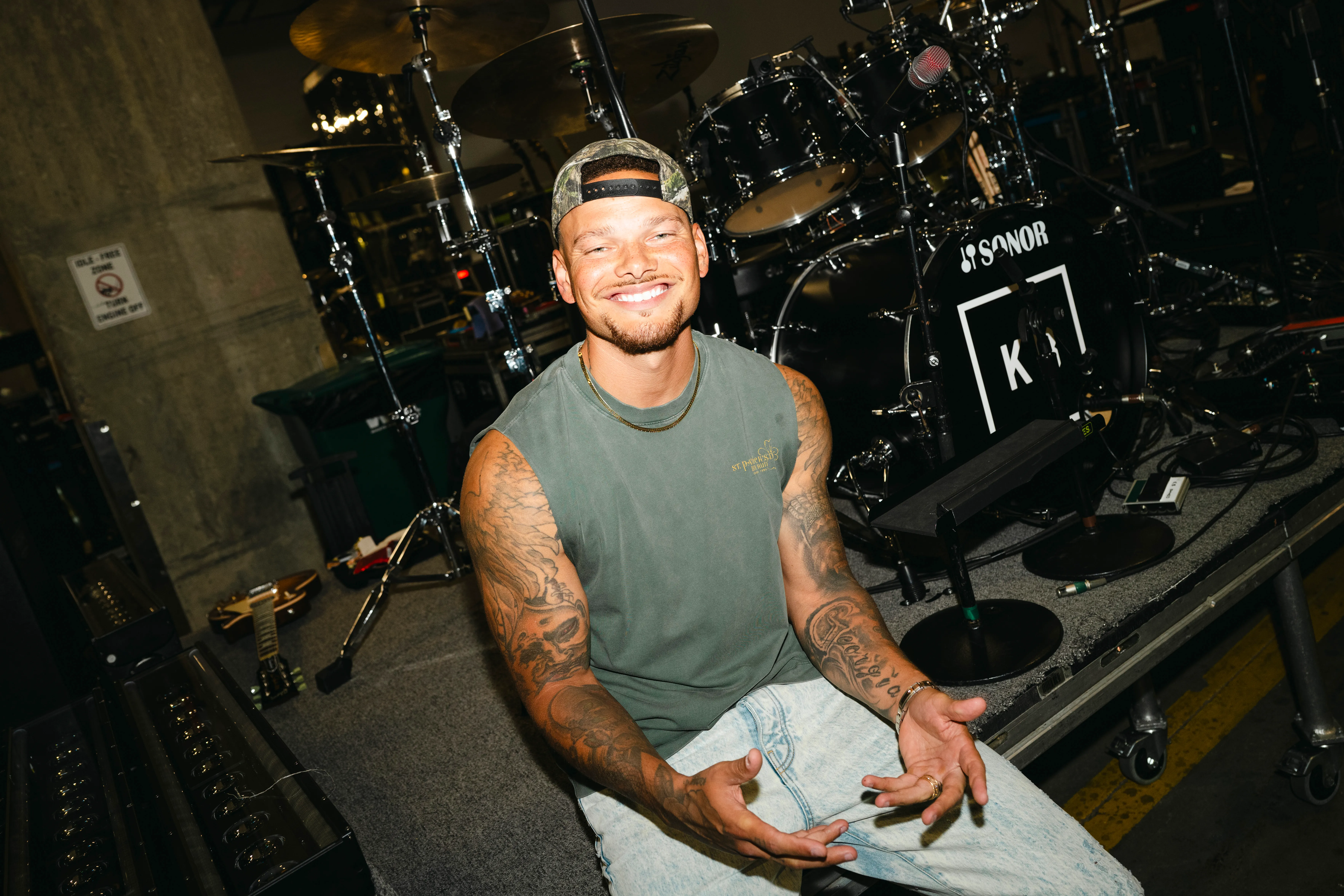 Country singer Kane Brown smiles with a drum kit behind him.