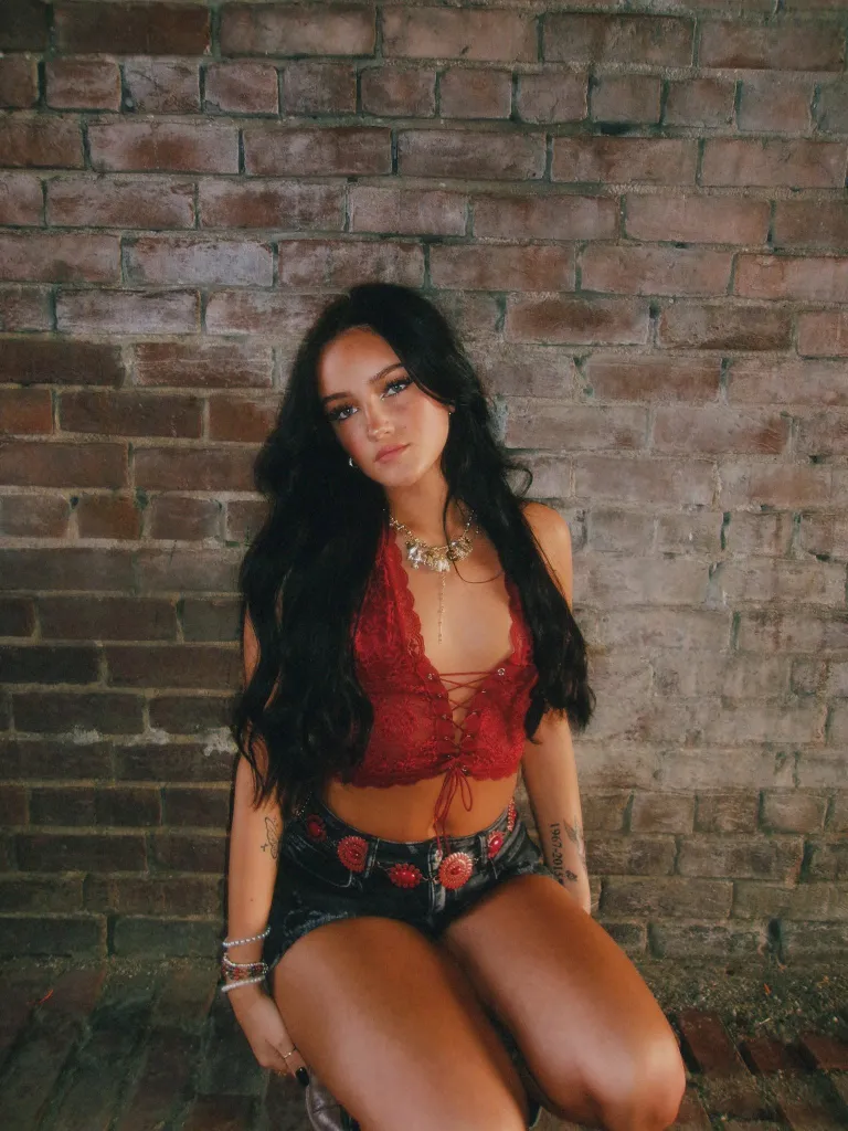 Marly Kinney, a young woman with long dark hair, wearing a red lace top and denim shorts, kneels against a brick wall.