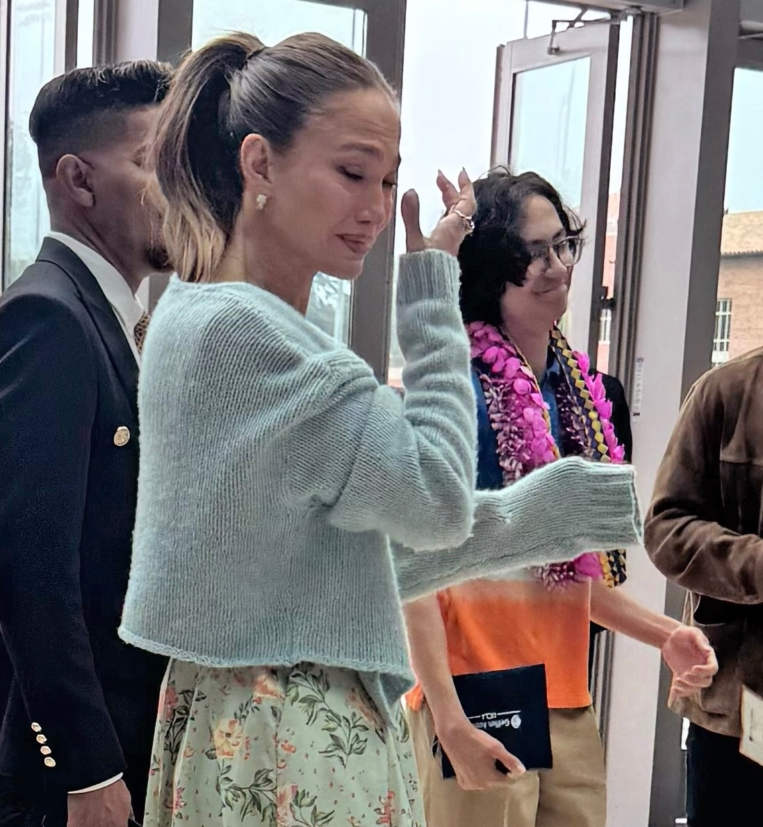 Jennifer Lopez, wearing a floral dress, smiles for a photo with a group of people, with one person holding a diploma.