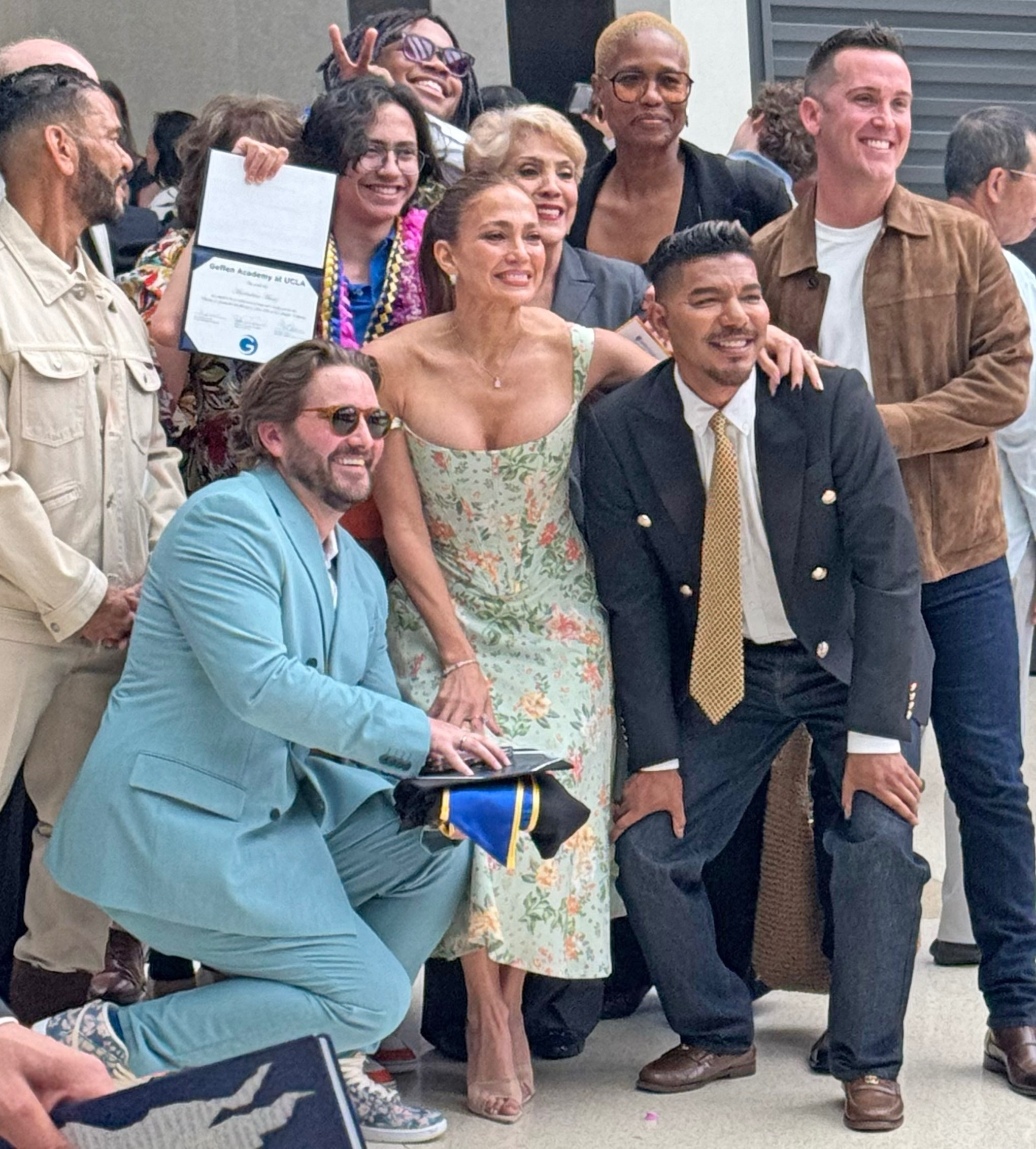 Jennifer Lopez, wearing a floral dress, smiles for a photo with a group of people, with one person holding a diploma.