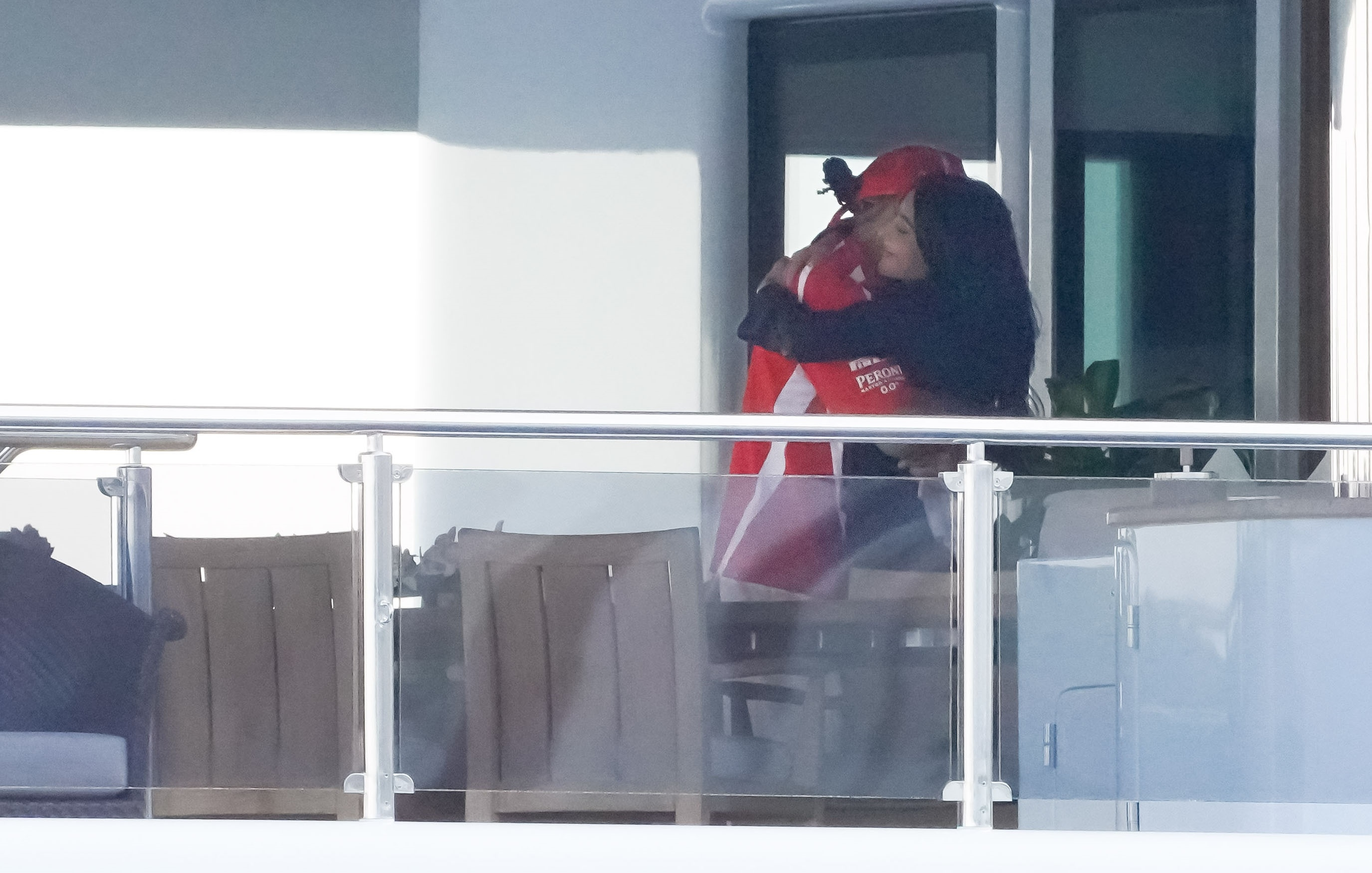 Kim Kardashian in a black long-sleeve top hugging Lewis Hamilton wearing a red baseball cap and red team polo shirt with logo patches.