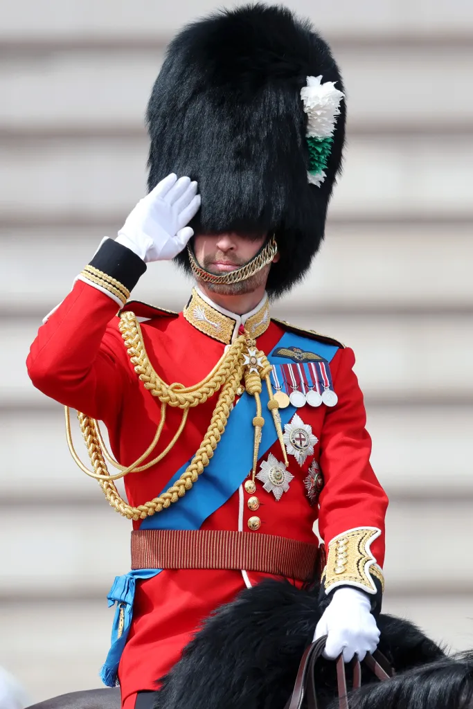 Prince William saluting while wearing a red military uniform with a tall bearskin hat, gold braids, and medals.