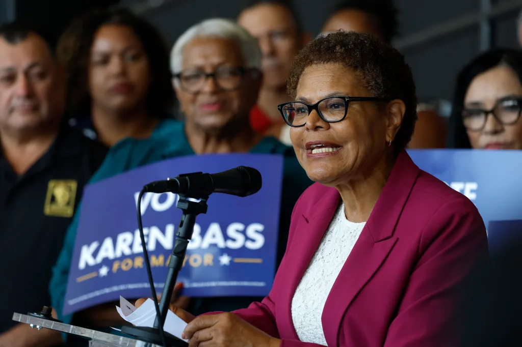 Karen Bass speaking at a campaign rally, with supporters and campaign signs in the background.