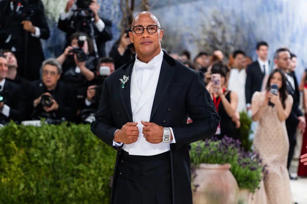 Dwayne Johnson in a black tuxedo jacket with white bow tie and vest, adorned with a green and silver brooch, at the Met Gala.