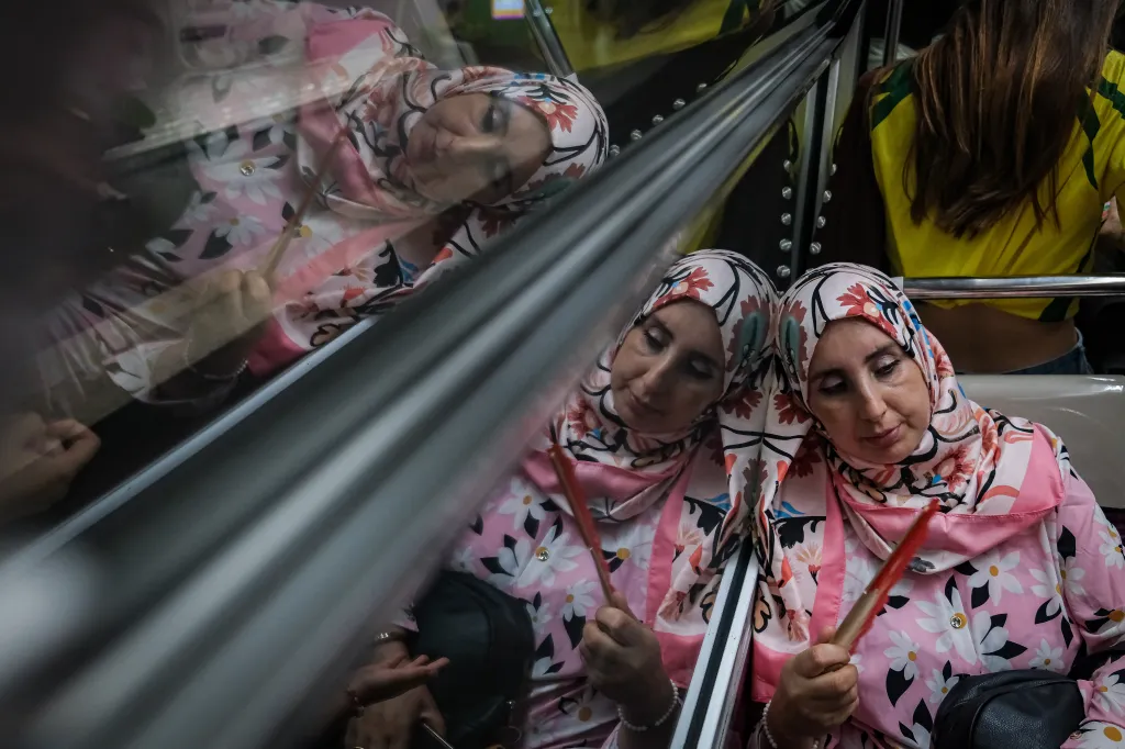 A passenger wearing a pink headscarf and shirt with white flowers holds a hand fan while riding a Paris Metro train.