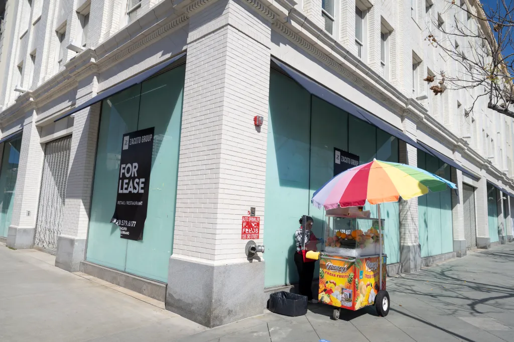 A fruit vendor with a rainbow umbrella stand in front of a vacant retail space on 3rd Street Promenade in Santa Monica, California.