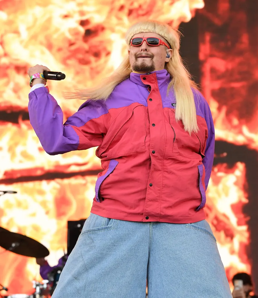 Oliver Tree performing in a red and purple jacket, baggy blue jeans, and red sunglasses, holding a microphone against a backdrop of fire.