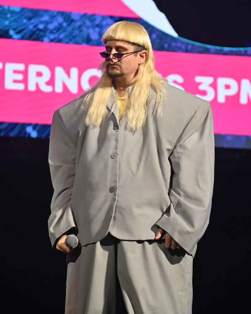 Oliver Tree performing in a light gray suit with a yellow shirt, sunglasses, and blonde bowl cut.