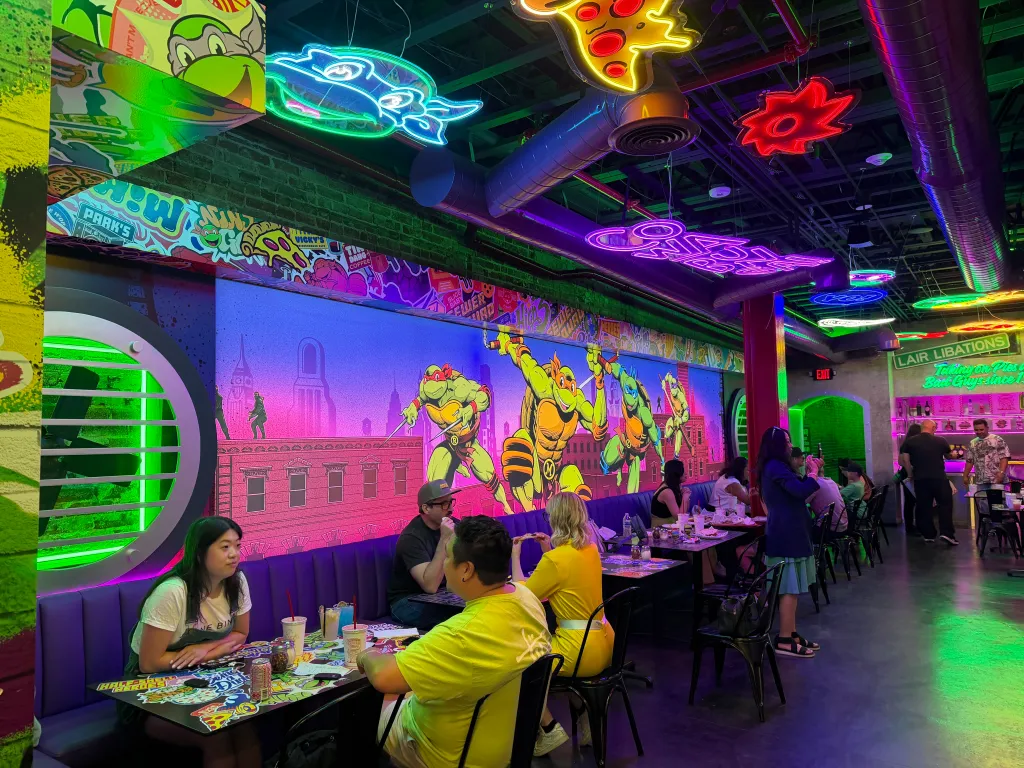 The interior of the Teenage Mutant Ninja Turtles Pizzeria at the Promenade, featuring colorful neon signs, cartoon murals of the Ninja Turtles, and people dining at tables with purple booths.
