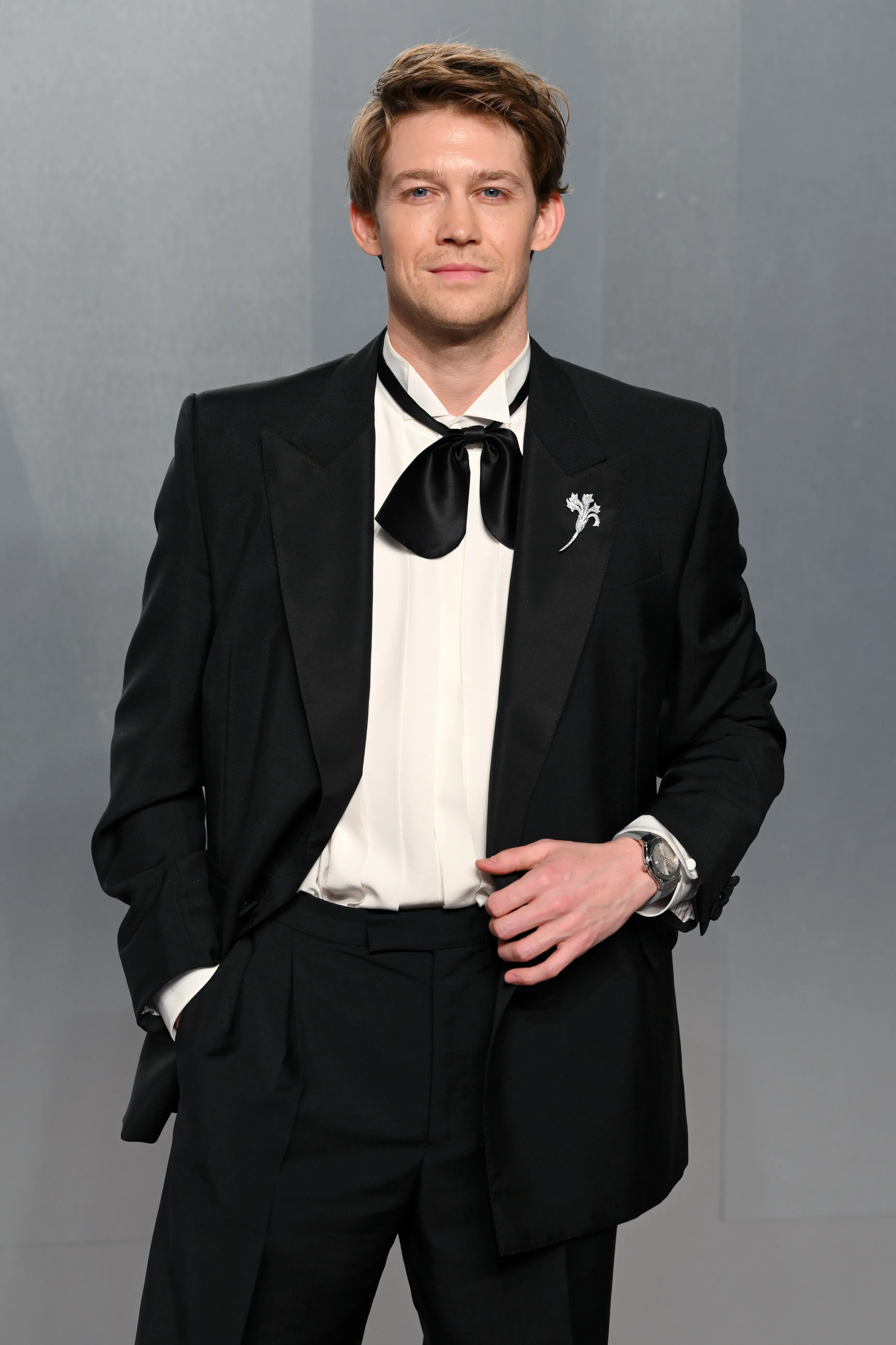 Joe Alwyn poses in a black suit with a large black bow tie and a silver brooch.