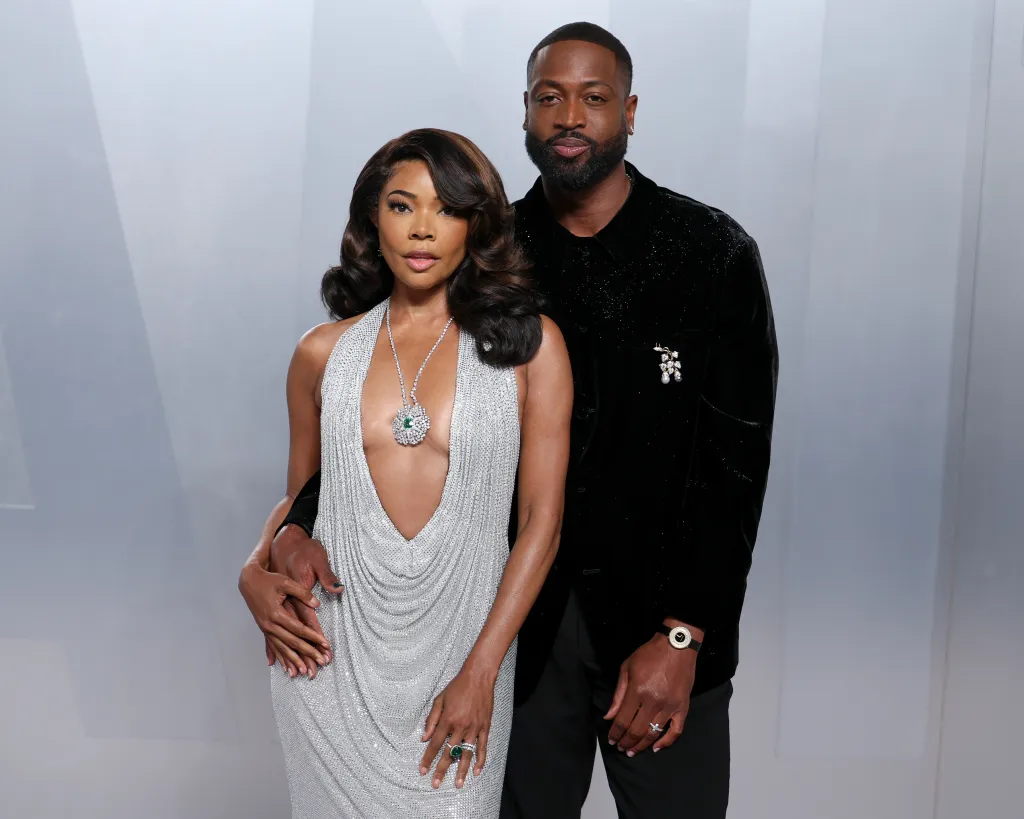 Gabrielle Union in a silver dress with a deep V-neck and a large emerald and diamond necklace, and Dwyane Wade in a black velvet jacket and black trousers, at the 2026 Vanity Fair Oscar Party.