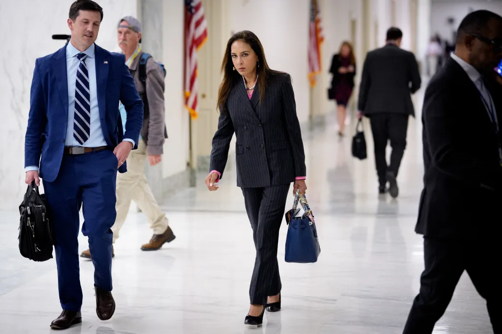 Assistant Attorney General for Civil Rights Harmeet Dhillon in a pinstripe suit, holding a blue handbag, walking past a man in a blue suit with a striped tie, through a brightly lit hallway.