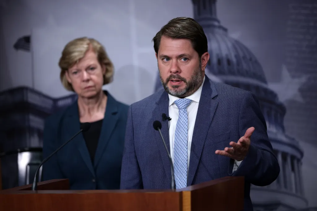 Sen. Ruben Gallego speaking at a podium with Sen. Tammy Baldwin listening in the background.