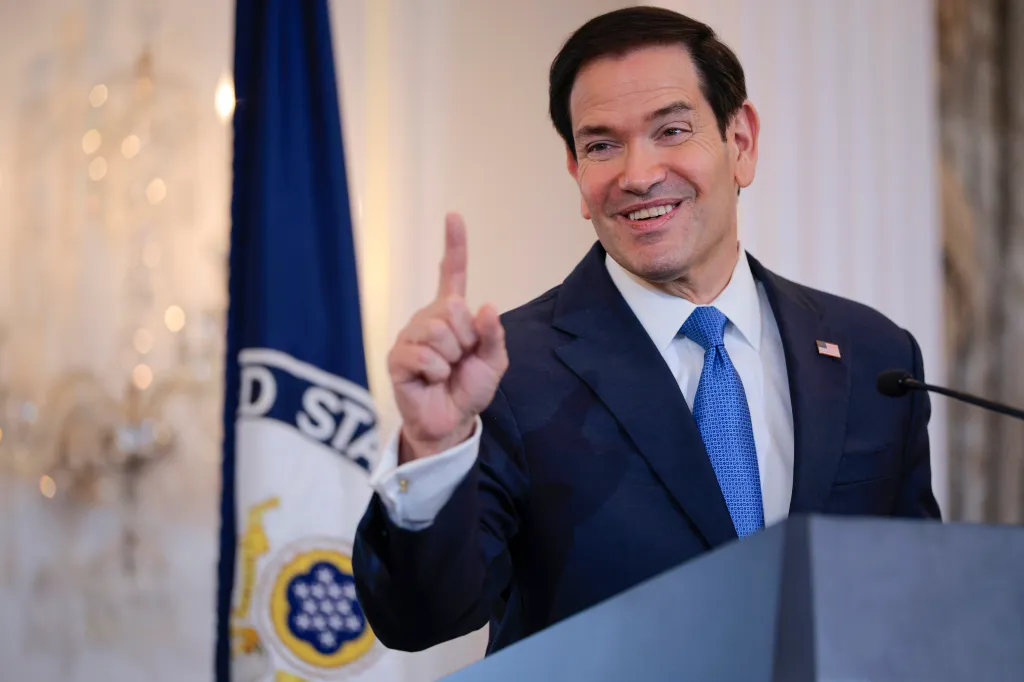 Marco Rubio smiling and pointing while speaking at a podium with a US State Department flag behind him.