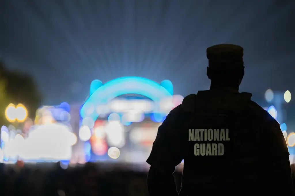 A National Guard member facing away from the camera, silhouetted against a blurred background of colorful lights, overseeing the UFC Freedom 250 crowd near the White House.