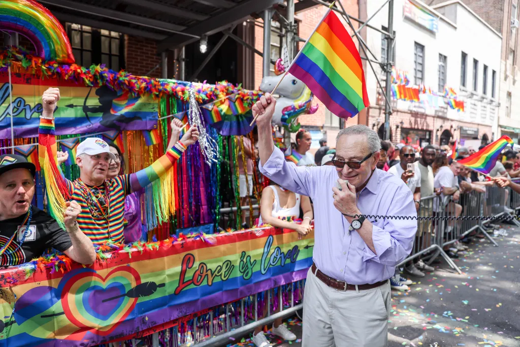 A man in a light purple shirt and sunglasses holds a small rainbow flag in one hand and a microphone in the other at the NYC Pride March.