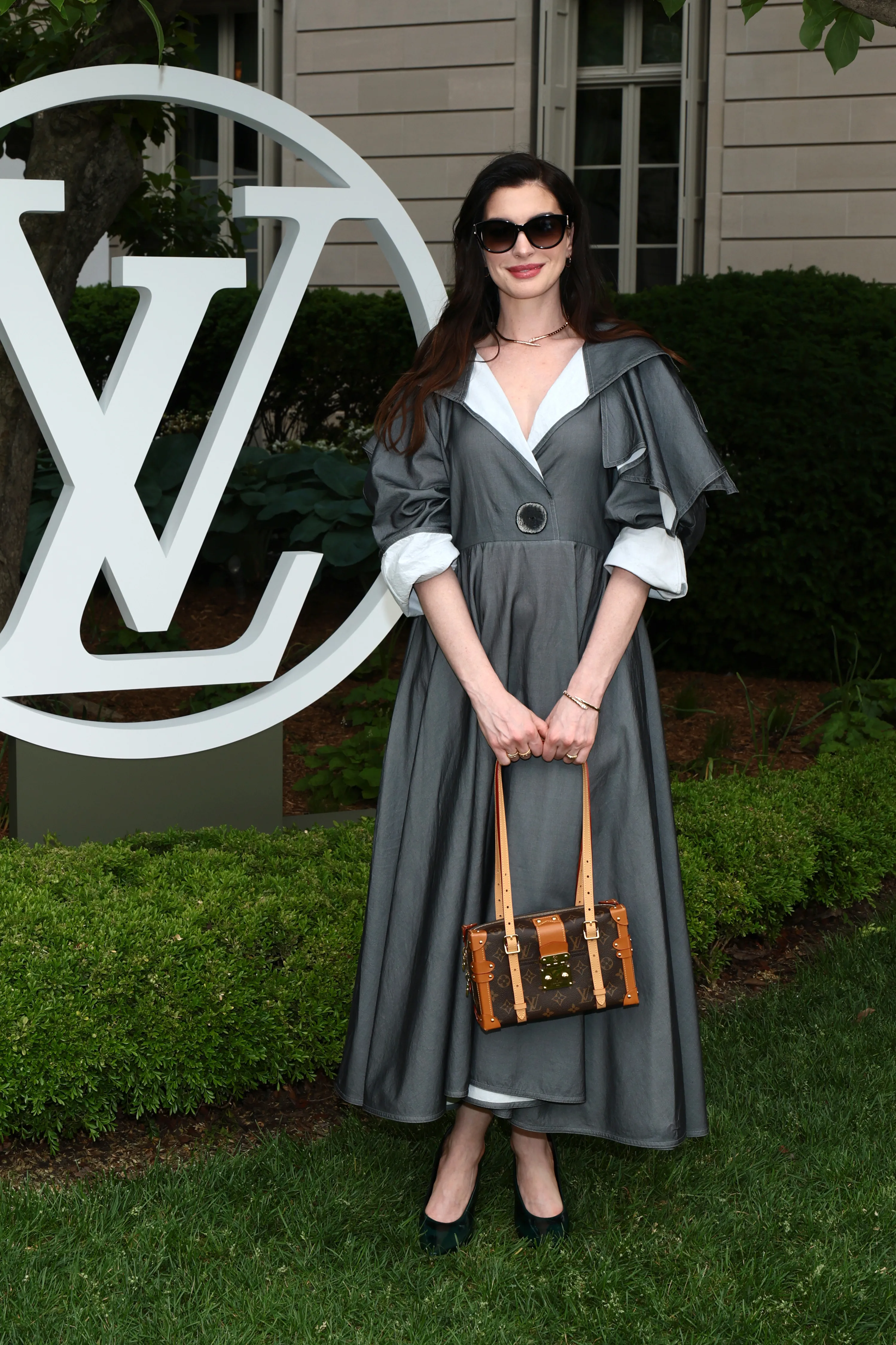 Anne Hathaway in a baggy grey dress and sunglasses holding a Louis Vuitton bag at the Louis Vuitton Cruise 2027 show.