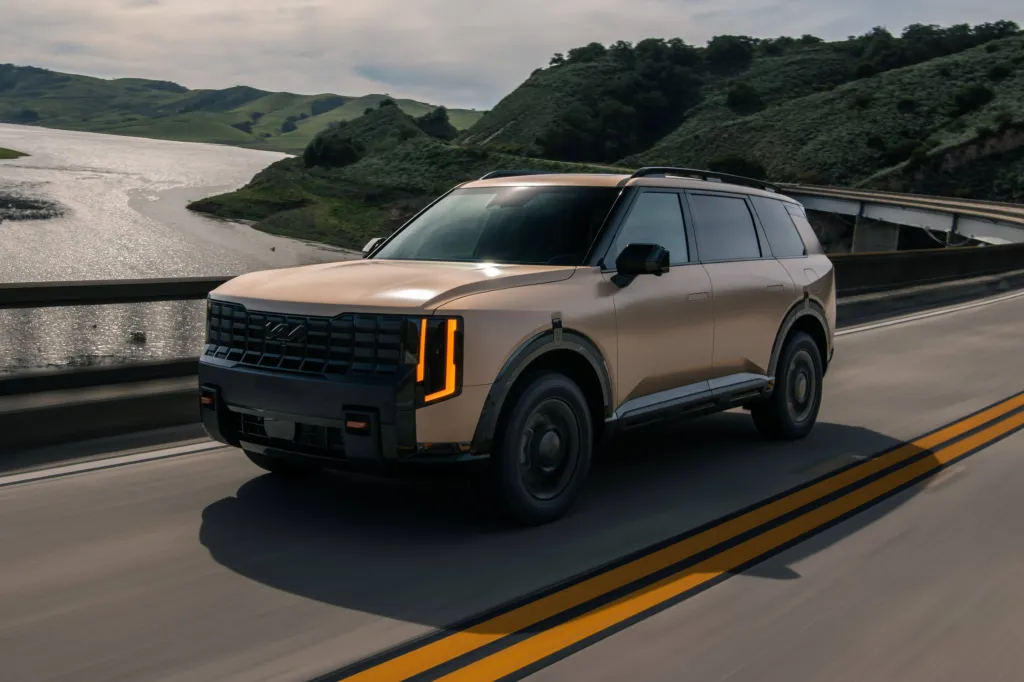A 2027 Kia Telluride, a three-row SUV, drives on a road next to a lake with green hills in the background.