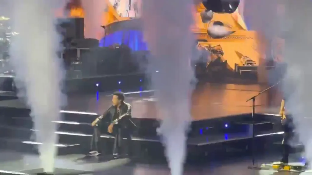 Lionel Richie sitting on stage in a black jacket and pants, surrounded by smoke effects.