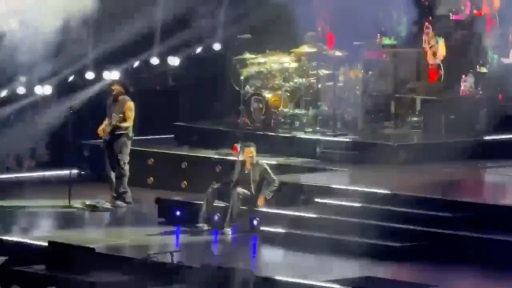 Lionel Richie crouching on stage during a performance, with a bandmate and drum kit visible in the background.