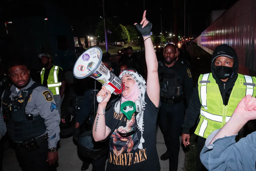Immigration advocate protesting at night with a megaphone while officers stand by.