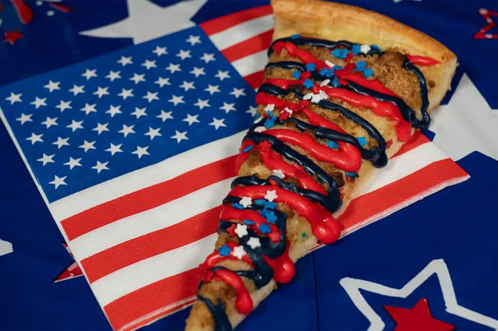 Slice of patriotic dessert pizza with red, white, and blue icing and sprinkles, resting on a folded American flag napkin.