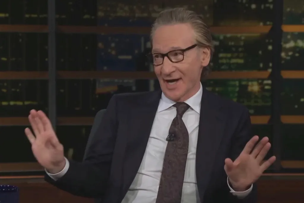 Comedian Bill Maher speaking with his hands raised in front of a dark window.