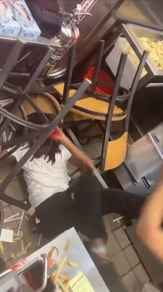 A person injured on the floor of a fast food restaurant amidst overturned chairs and scattered food.