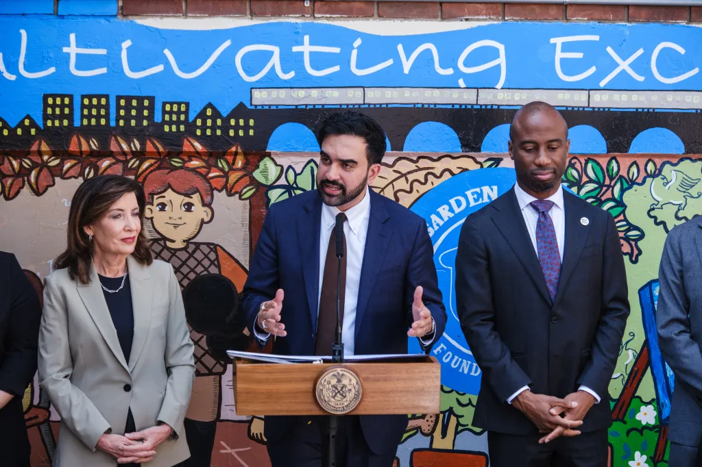 New York State Assemblyman Zohran Mamdani speaking from a podium between New York Governor Kathy Hochul and a man in a suit, in front of a colorful mural.