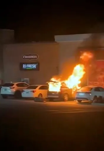 A vehicle engulfed in flames in a parking lot at night.