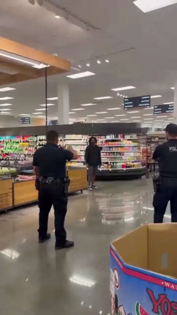 A man with long curly hair and a beard, wearing a black shirt and shorts, standing in a Target aisle surrounded by three police officers.