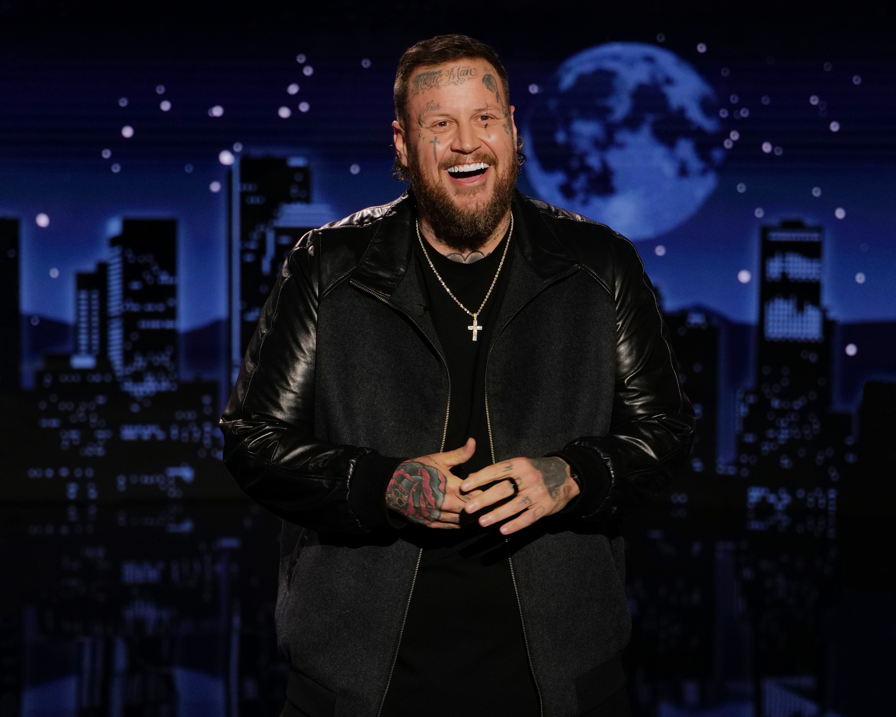 Jelly Roll smiling in a black jacket against a dark blue city skyline with a full moon.
