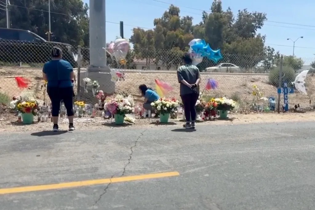 Three people standing at a roadside memorial with flowers, candles, and balloons.