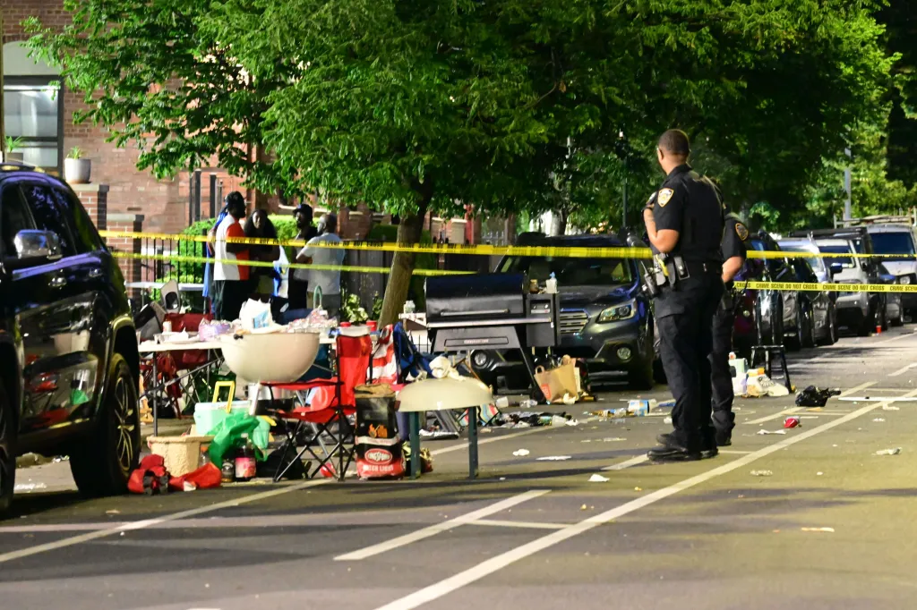 NYPD Crime Scene Unit investigating the fatal shooting of Emanuel Spencer at 101 Lincoln Road, Brooklyn, with yellow caution tape, debris on the street, and two police officers.