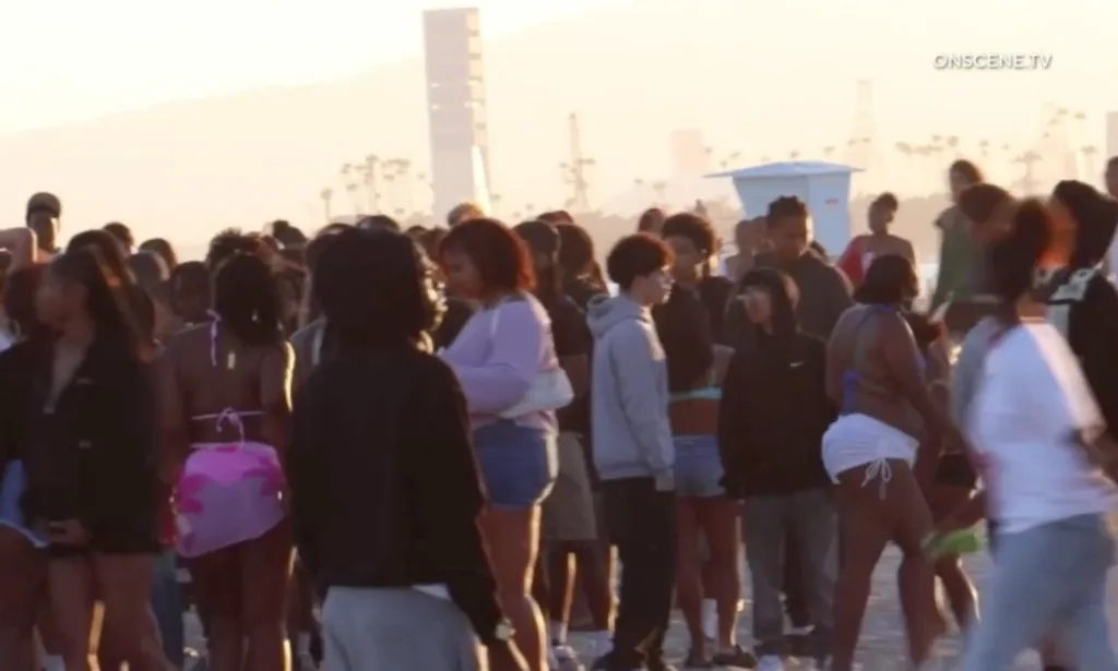 A large crowd of young people gathered on a beach, with buildings and palm trees in the background.