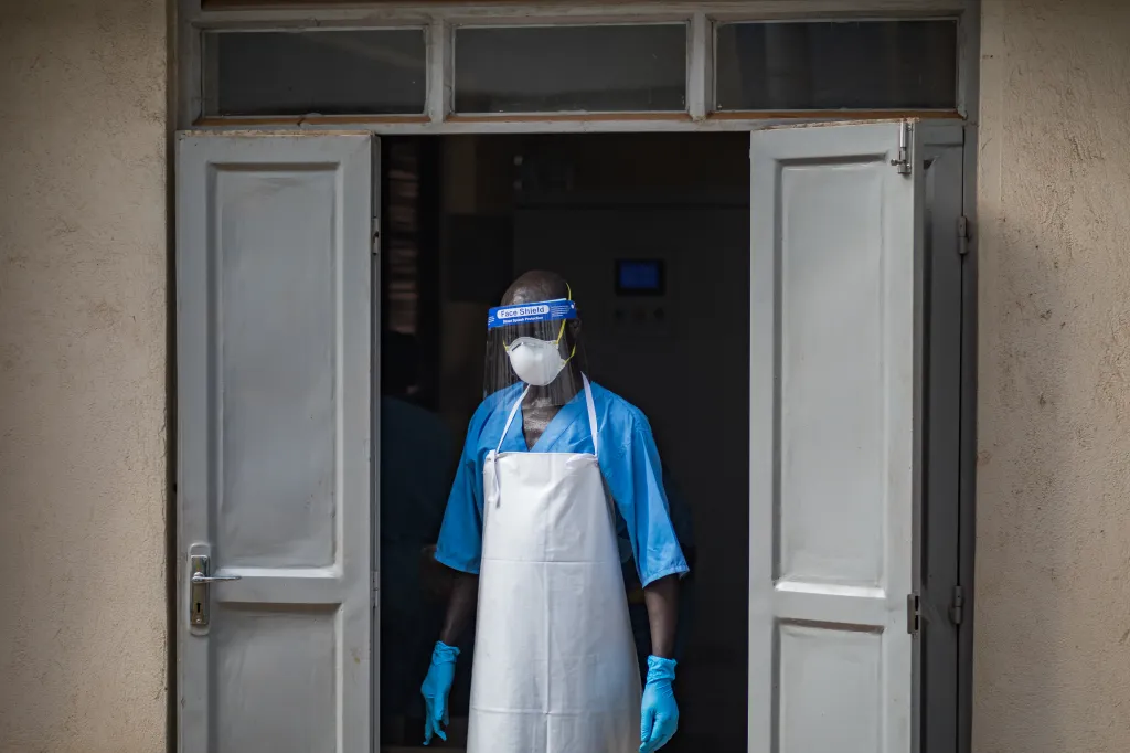A health worker wearing a face mask, face shield, blue scrubs, white apron, and blue gloves stands in a doorway.