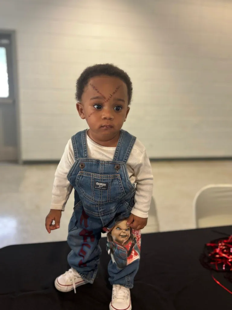 A baby dressed as Chucky from Child's Play, with overalls, a white long-sleeve shirt, makeup scars on the face, and Chucky printed on the pants.