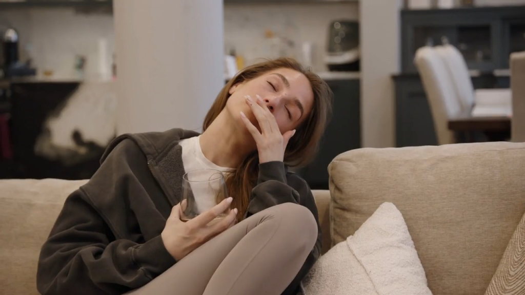 A woman on a couch holding a glass, her head back and hand to her mouth, appearing unwell.