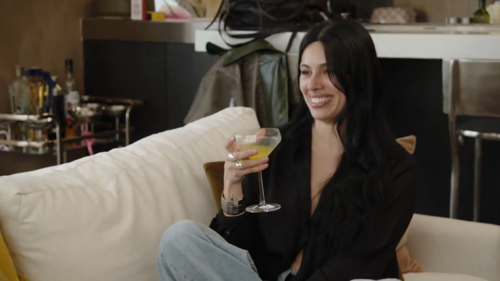 A smiling woman with long dark hair, wearing a black top and jeans, holding a cocktail.