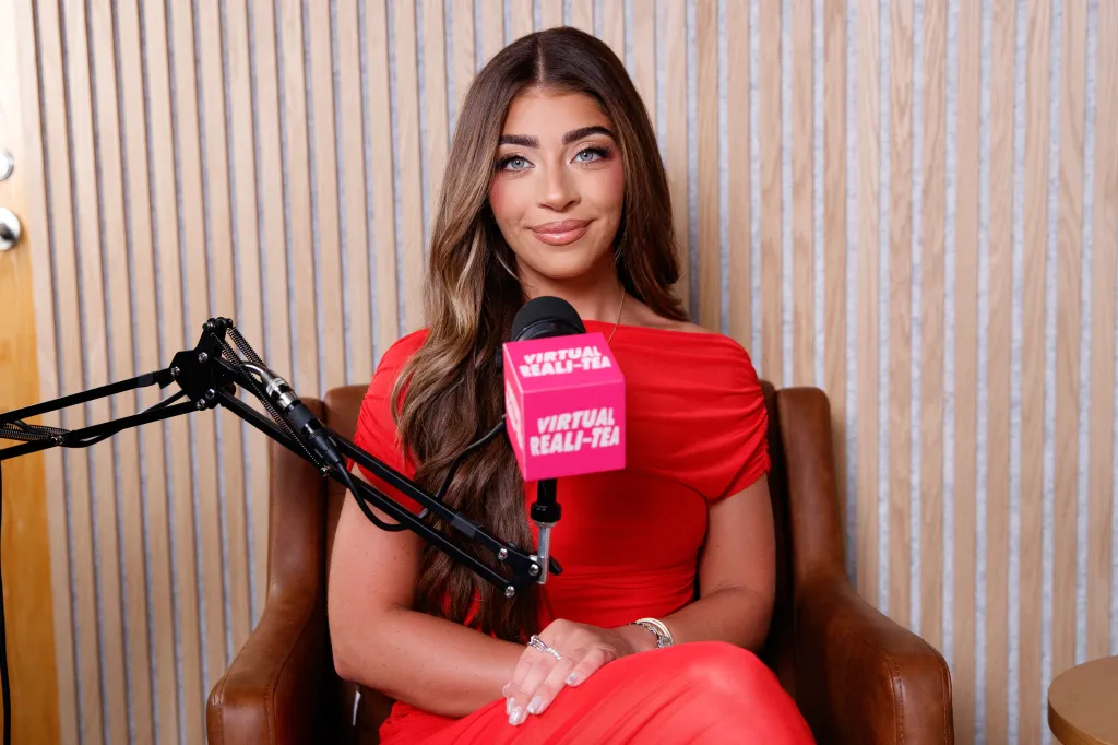 Gia Giudice sitting in a brown leather chair, wearing a red dress, behind a microphone with a pink box that says