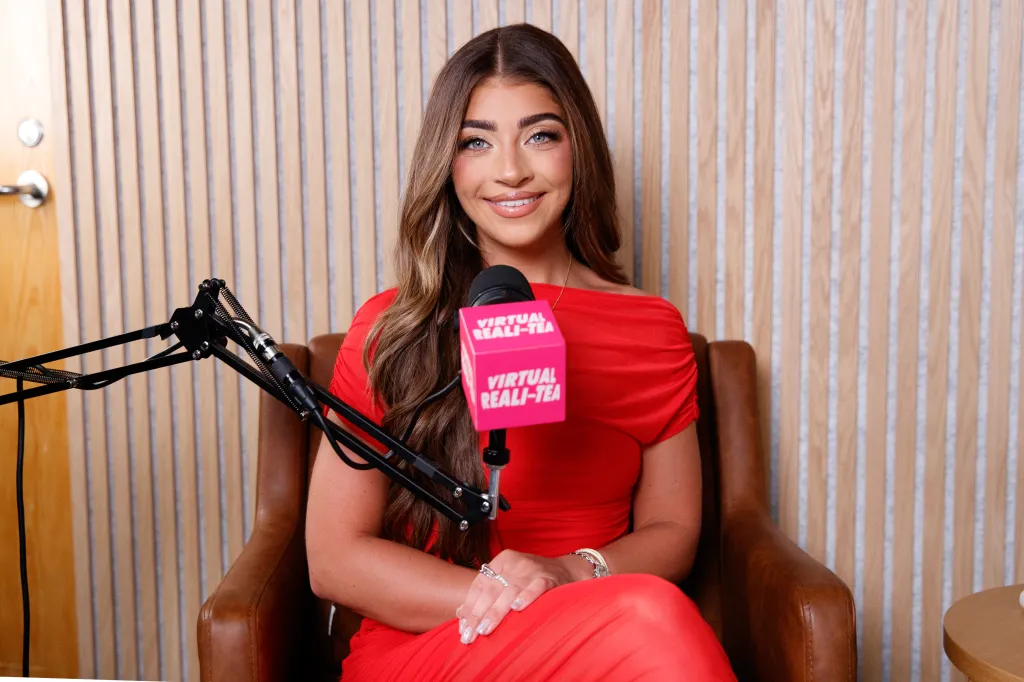 Gia Giudice sits in a brown leather chair, wearing a red off-the-shoulder dress, smiling at the camera with a