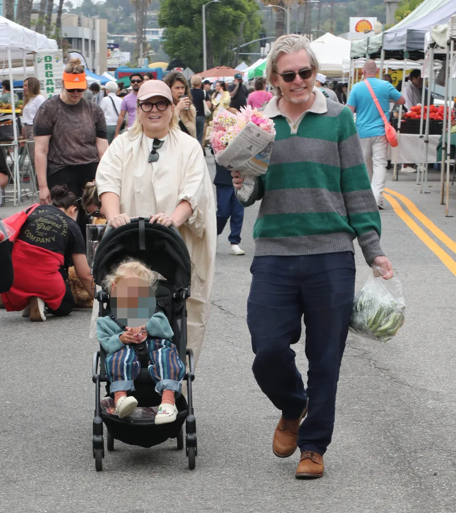 Sia pushing a child in a stroller at a farmers market, while a man next to her carries flowers and a bag of produce.