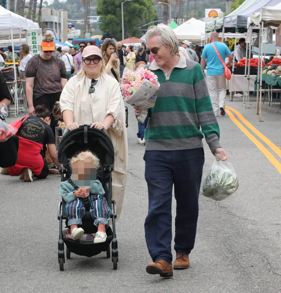 Sia pushing a child in a stroller at a farmers market, while a man carrying flowers and groceries walks beside her.