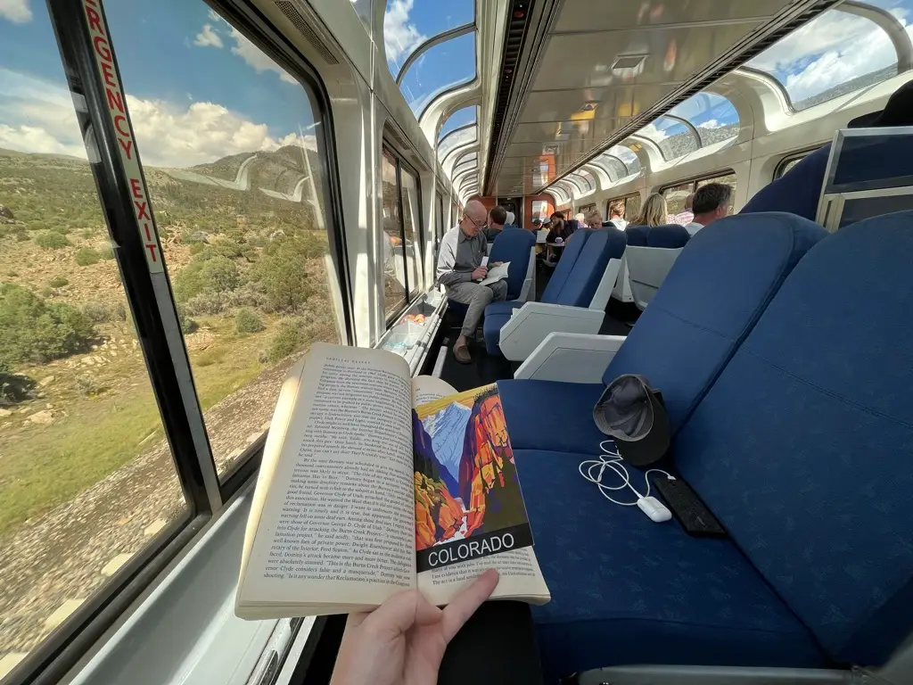 A person holding a book open with a Colorado bookmark inside while riding on an Amtrak train with grassy hills seen through the window to the left.