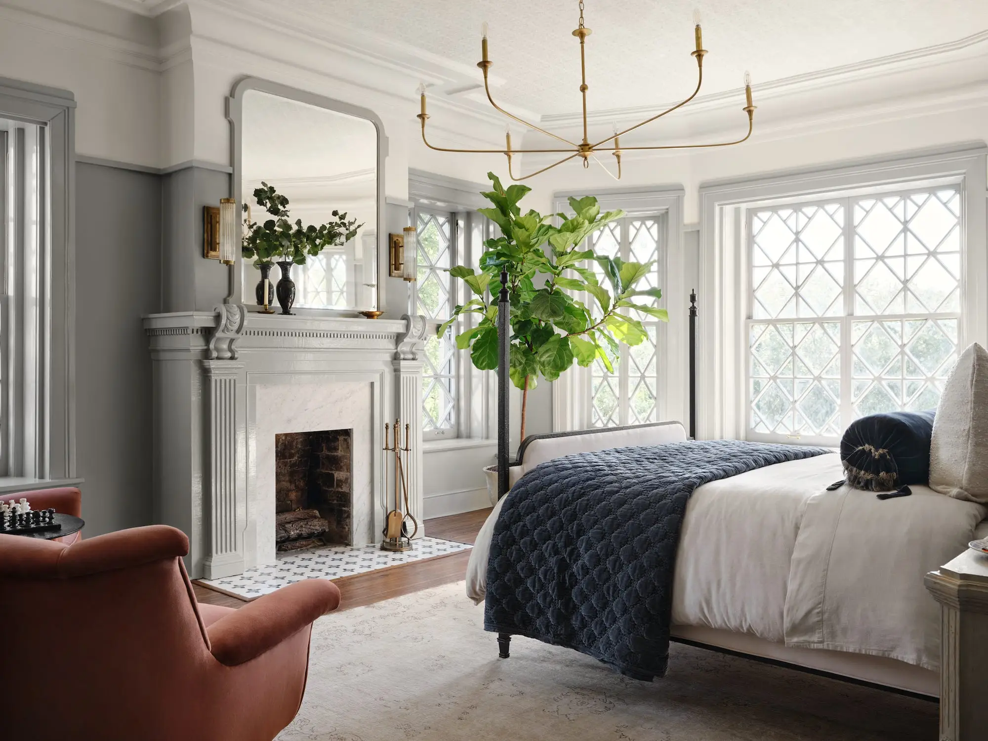 A bedroom with a white fireplace, big chandelier, and large windows.