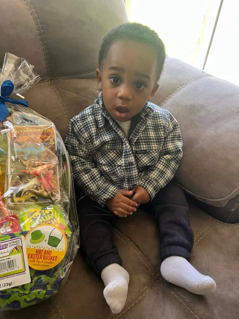 A young boy with short, curly hair sits on a couch next to a wrapped Easter basket filled with dinosaur toys, candy, and a hat.