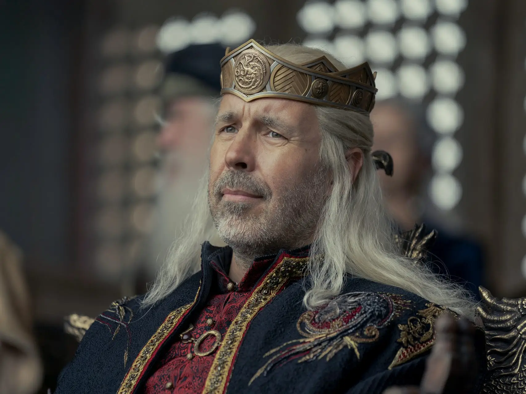 A man in his 50s with long silver hair and wearing a gold crown looks pensively out towards a crowd.