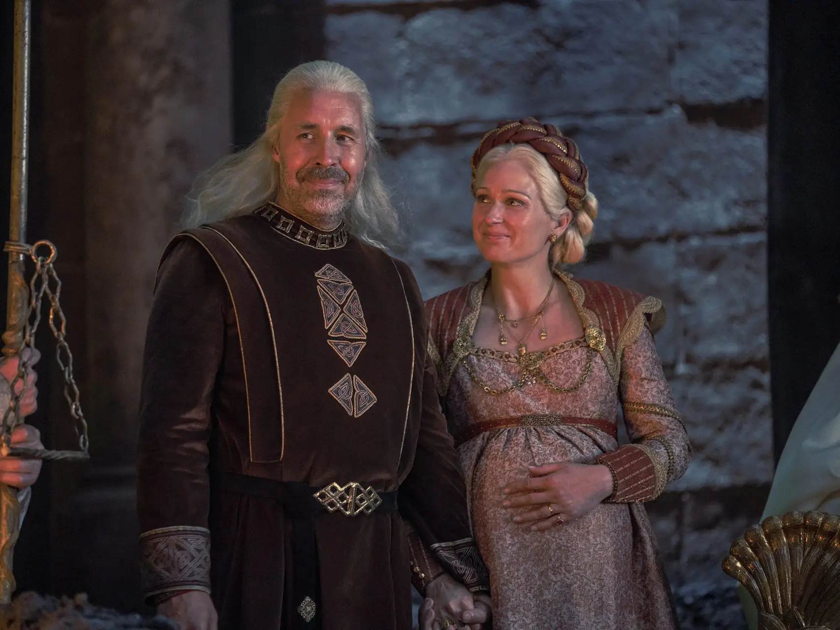 A man and woman with silver hair wearing medieval royalty garb stand lovingly next to each other.