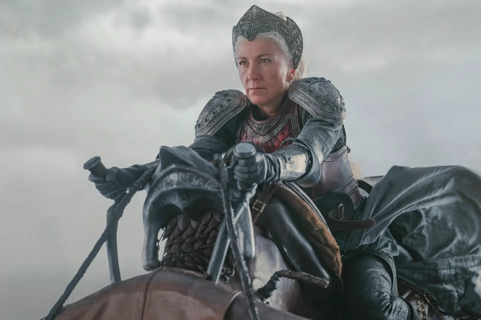 eve best as rhaenys targaryen in house of the dragon, dressed in full armor and a headpiece while clinging onto a dragon statue. she's flying through a grey sky, and her expression is resolute